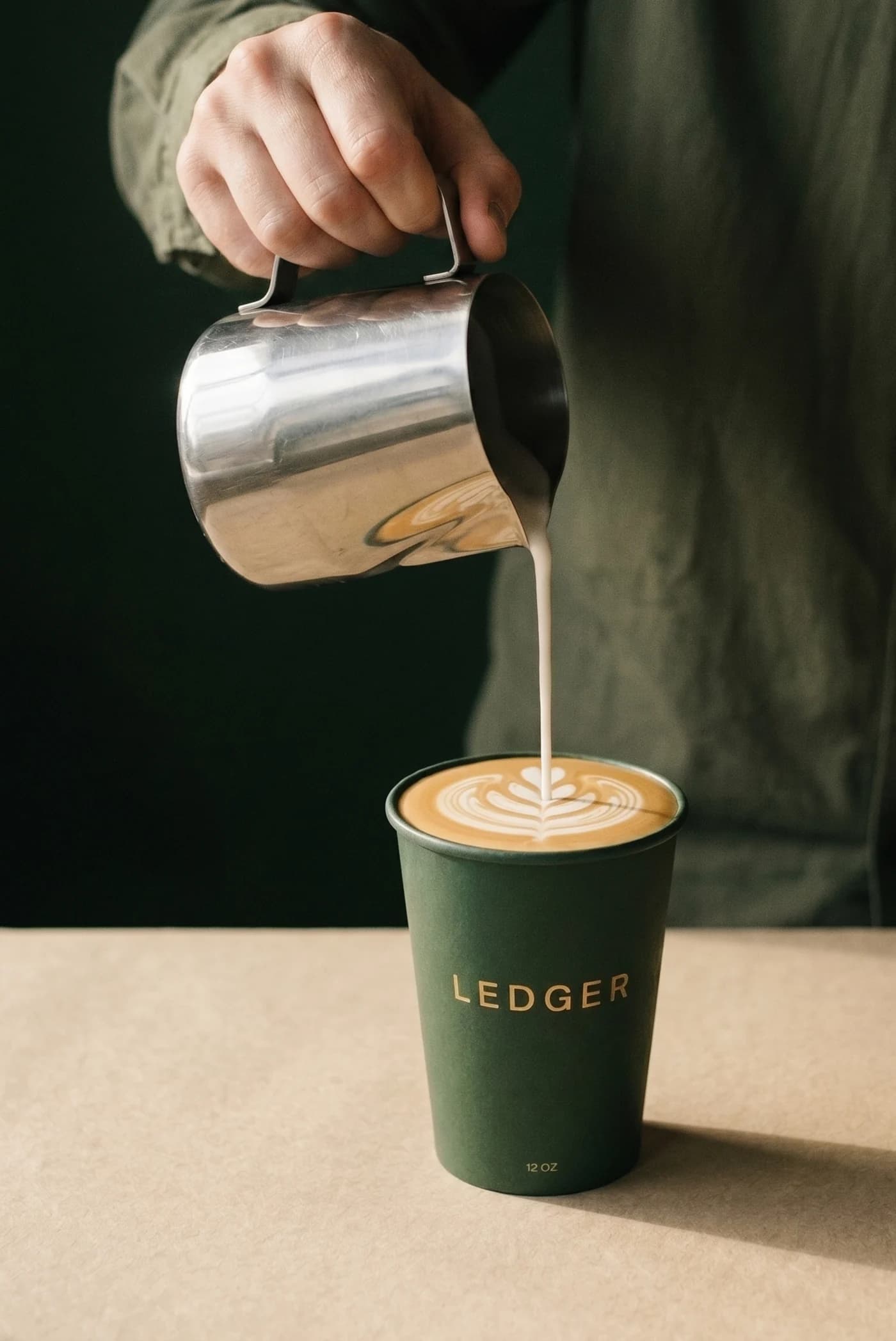 A barista pouring a rosetta into the bottle-green LEDGER cup